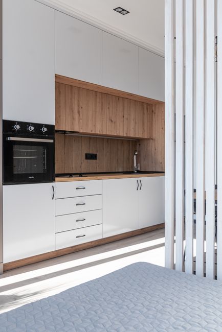 A modern kitchen with grey matte finish cabinets and a light wood floor, featuring a built-in stainless steel oven, a black range hood, and a black kitchen sink with a curved faucet, set beneath large