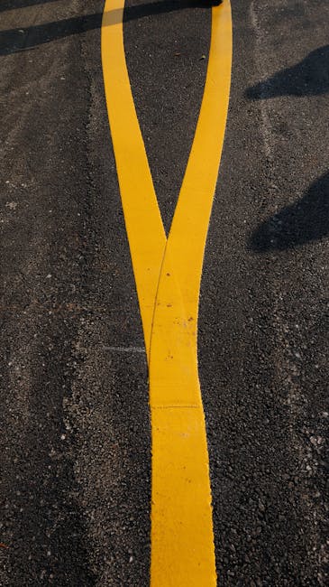 Close-up view of a parking lot surface featuring a yellow line marking that is slightly cracked and worn, painted on dark asphalt. The line curves moderately and appears freshly painted with vibrant color, though some dirt and wear are visible, indicating regular use. Shadows cast by nearby objects or structures fall across parts of the surface, adding depth to the scene. This image emphasizes surface marking maintenance relevant to building exterior or pavement cleaning services, as provided by Cleaners Harringay, which specializes in surface cleaning, deep cleaning, and sanitisation for residential and commercial properties near Green Lanes in Harringay.