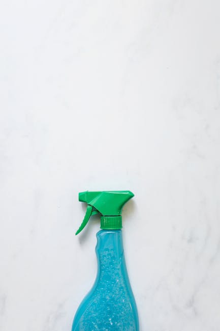 A close-up image of a blue plastic spray bottle with a green trigger and nozzle, positioned against a plain white wall. The surface of the wall appears to have some minor smudges or dirt marks, suggesting it may require surface cleaning or sanitisation. The lighting is neutral and evenly distributed, highlighting the clean and simple presentation of the cleaning product. This image likely illustrates cleaning tools used for domestic or commercial cleaning tasks, aligning with the services offered by Cleaners Harringay in their end of tenancy cleaning guide for Harringay as part of their comprehensive surface and surface area cleaning solutions.