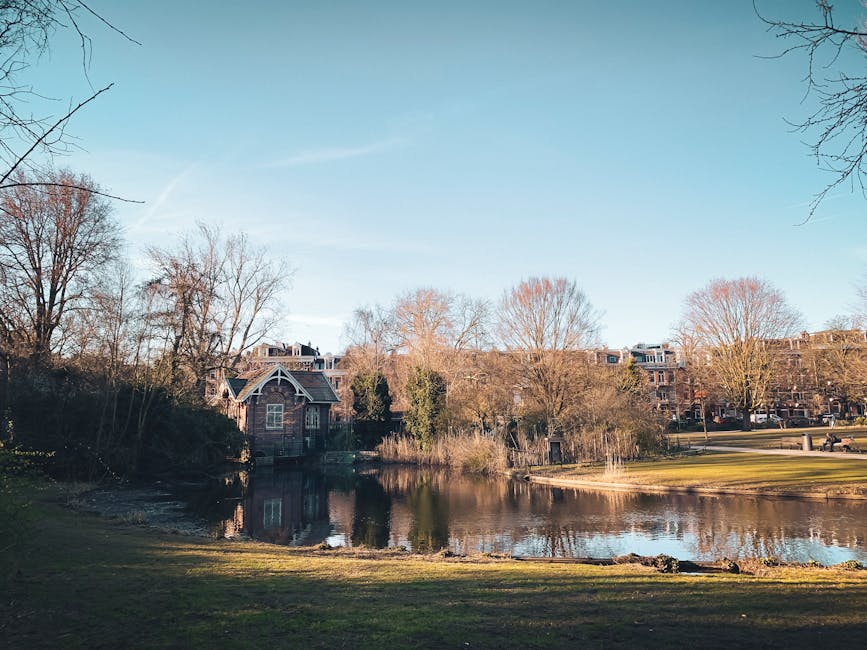 Photograph of a peaceful residential area featuring a small pond surrounded by leafless trees and well-maintained grass. Behind the pond, there are several brick and stone houses with pitched roofs, l